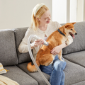 Woman on a couch using a pet grooming vacuum with clippers to trim a calm Shiba Inu held in her lap